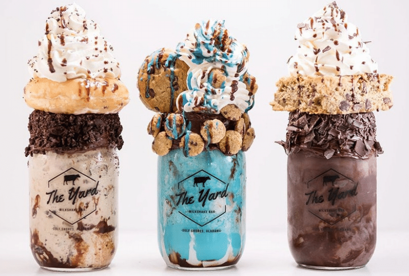 Three milkshakes in glass mason jars from The Yard. From left to right, one vanilla with a donut and whipped cream, one blue with cookies and whipped cream and lastly one chocolate with a cookie and whipped cream.