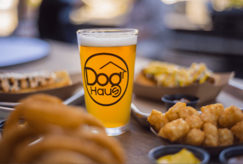Dog Haus tall glass beer with food in dishes surrounding the glass